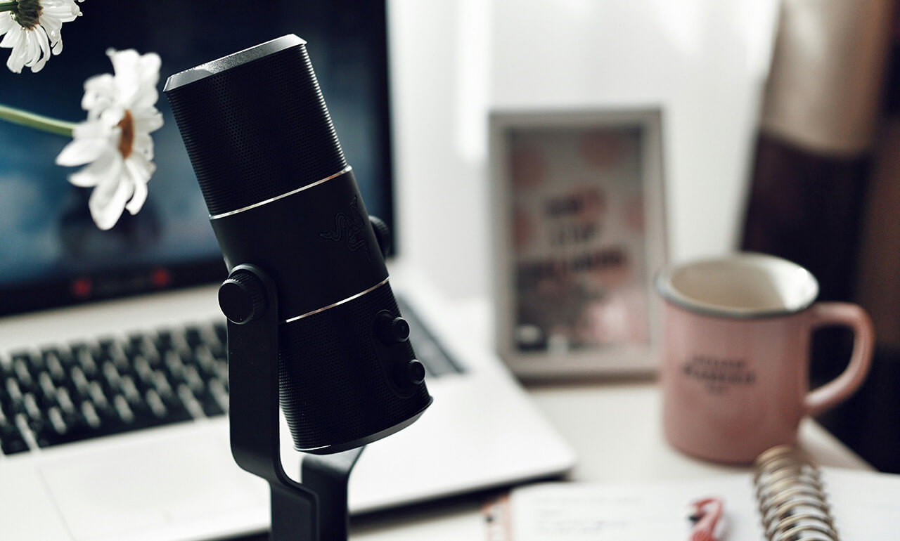 A desk with a microphone visible in the foreground. In the background, there is a laptop, a coffee mug and a notepad with a pen.